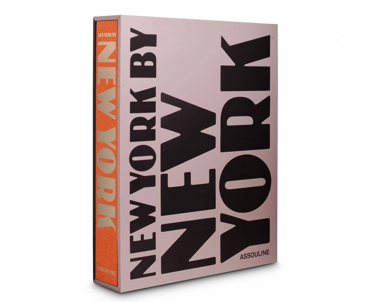 A large hardcover photography book, "New York By New York" by Assouline, features bold black text on a light pink cover with an orange spine. The book stands upright and is slightly angled to the right.