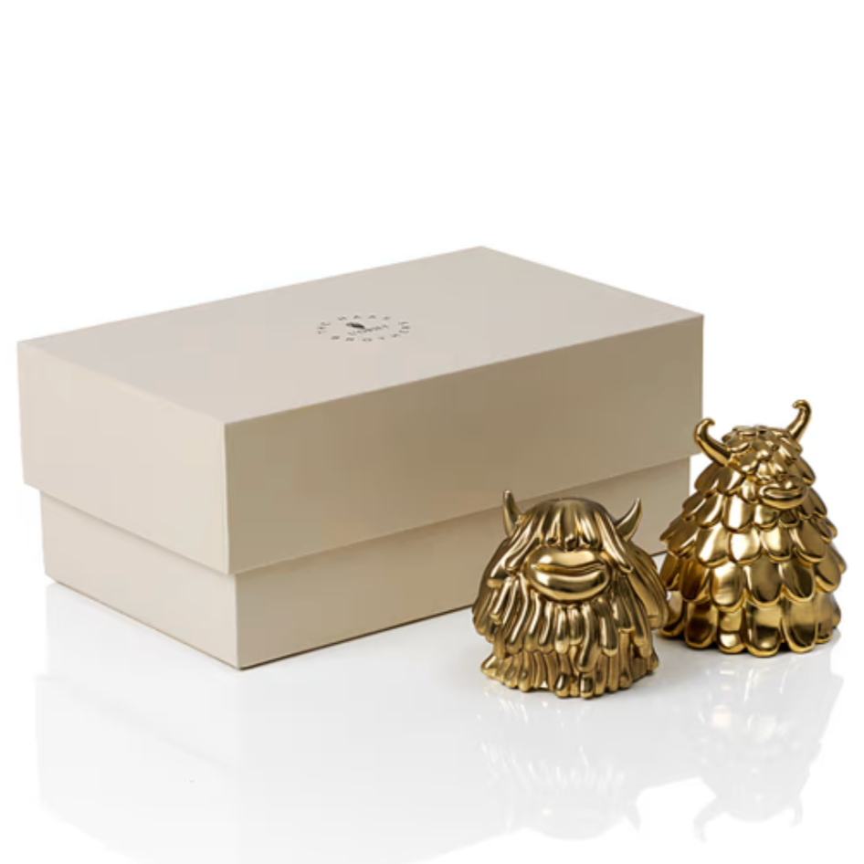 A cream-colored gift box is displayed beside two small, shiny gold Niki & Simon Salt & Pepper shakers by Haas Brothers for L’Objet—whimsical, monster-shaped brass pieces with textured bodies and quirky horns.