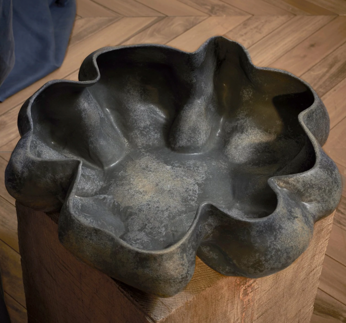 The Timna Bowl Blue by L’Objet—a dark, irregularly shaped metal vessel with wavy, petal-like edges—rests atop a wooden block on a light wood floor, its sculptural form evoking treasures unearthed from ancient times.