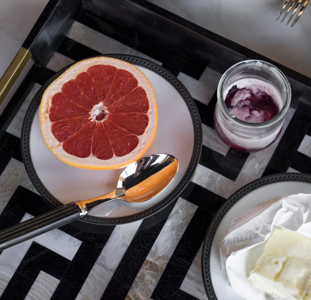 A halved grapefruit on a L'Objet Soie Tressée Black plate with a spoon, beside yogurt with berry swirl and wrapped cheese, all displayed on a black and white geometric tray.