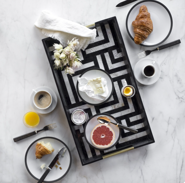 Breakfast is elegantly served on L'Objet Soie Tressée Black Dinnerware, featuring coffee, orange juice, croissants, grapefruit, cheese, egg, and yogurt on luxury porcelain plates atop marble. A black tray holds flowers and neatly arranged dishes from above.
