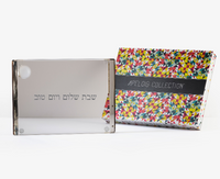 A Personalized Solid Acrylic Challah Board by Apeloig, etched with Hebrew text, stands beside a colorful geometric-patterned Apeloig Collection box featuring a black band.