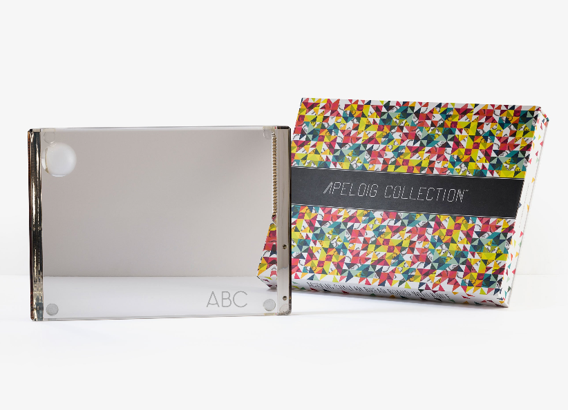 A clear rectangular Personalized Solid Acrylic Challah Board by Apeloig, engraved with ABC, sits in front of a colorful patterned box labeled Apeloig on a white background.