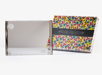A Personalized Solid Acrylic Challah Board by Apeloig, featuring initials in the corner, is displayed beside a vibrant patterned box labeled “APELOIG COLLECTION” on a white background.