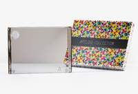 A clear, rectangular Personalized Solid Acrylic Challah Board by Apeloig with a visible spine stands before a colorful patterned box featuring the Apeloig name on a black band.