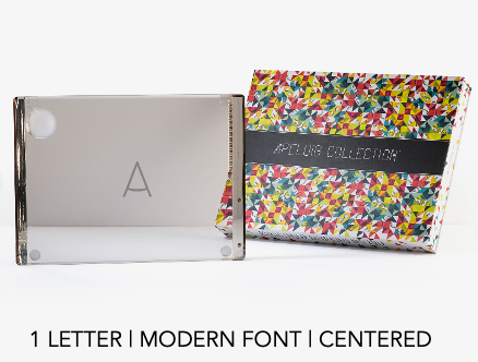 A glass block with the letter A in a modern, centered font stands next to a colorful, patterned Apeloig box. Text below reads: 1 letter | modern font | centered—an ideal accent for your Personalized Solid Acrylic Challah Board.