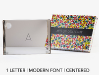 A glass block with the letter A in a modern, centered font stands next to a colorful, patterned Apeloig box. Text below reads: 1 letter | modern font | centered—an ideal accent for your Personalized Solid Acrylic Challah Board.