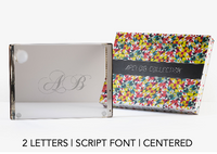 A Personalized Solid Acrylic Challah Board by Apeloig, engraved with script letters A B, is shown beside a colorful geometric Apeloig box. Text reads: 2 LETTERS | SCRIPT FONT | CENTERED.