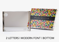 A personalized solid acrylic challah board by Apeloig, featuring two modern font letters at the bottom, comes in a colorful patterned Apeloig Collection gift box.