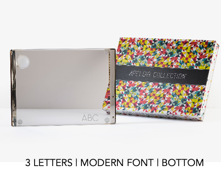 A rectangular, mirrored Personalized Solid Acrylic Challah Board by Apeloig featuring "ABC" in a modern font at the bottom corner, displayed beside a colorful patterned Apeloig box from the APELOVE COLLECTION.
