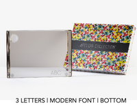 A rectangular, mirrored Personalized Solid Acrylic Challah Board by Apeloig featuring "ABC" in a modern font at the bottom corner, displayed beside a colorful patterned Apeloig box from the APELOVE COLLECTION.