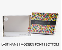 A rectangular, reflective Personalized Solid Acrylic Challah Board by Apeloig features a modern LASTNAME at the bottom and comes beside a colorful patterned Apeloig box. Text below: LAST NAME | MODERN FONT | BOTTOM.