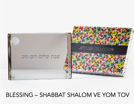 A Personalized Solid Acrylic Challah Board by Apeloig with Hebrew text stands beside a colorful, patterned Apeloig box. The English caption reads: BLESSING – SHABBAT SHALOM VE YOM TOV.