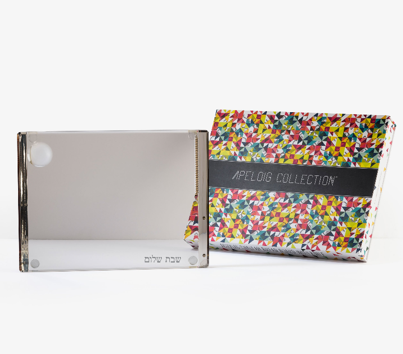 A Personalized Solid Acrylic Challah Board by Apeloig, featuring custom Hebrew engraving in the corner, is shown beside a vibrant patterned Apeloig box, both displayed on a white background.