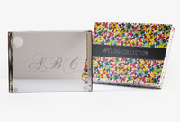 A clear acrylic photo frame engraved with ABC sits beside a colorful geometric box labeled Apeloig, reminiscent of the Personalized Solid Acrylic Challah Board from the Apeloig collection.