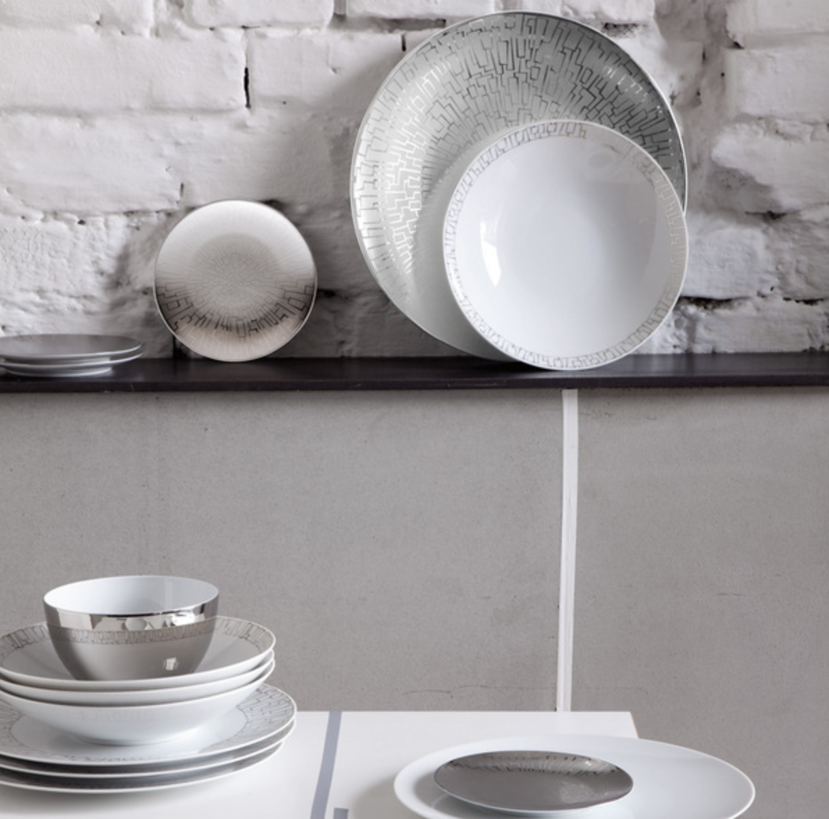 A modern table setting showcases stacked Rosenthal Skin Platinum TAC 02 Dinnerware—white porcelain with platinum accents and silver geometric patterns—on a white table before a brick wall and dark countertop with additional plates.