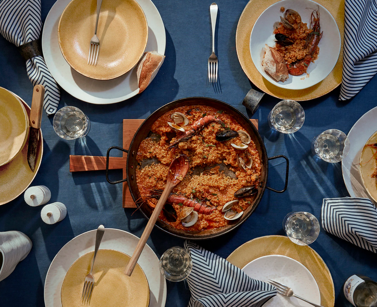 A pan of seafood paella is served on L'Objet Terra Leather Dinnerware, with yellow and white plates, utensils, napkins, wine glasses, and bread slices arranged atop a blue tablecloth.