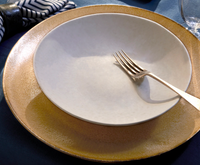 A Terra Leather Dinnerware bowl by L'Objet sits inside a gold charger plate, with a silver fork placed diagonally in the bowl. A blue-and-white striped napkin and a glass appear in the background.