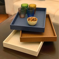 A set of three Bodrum Wicker Rectangle Trays in cream, brown, and blue are stacked on a dark surface. The top blue tray holds four textured glasses and a small bowl of nuts, with a beige sofa visible in the background.