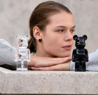A person rests their chin on their hands, looking at two Bear Brick art toys by Baccarat—one clear crystal, one black—displayed on a stone surface in front of them.