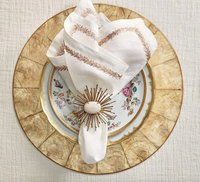 A decorative place setting features a gold-rimmed floral plate on a gold charger, with a folded white napkin embroidered with leaves and secured by the Kim Seybert Flare Napkin Ring Gold Set of 4, accented with a white stone center.