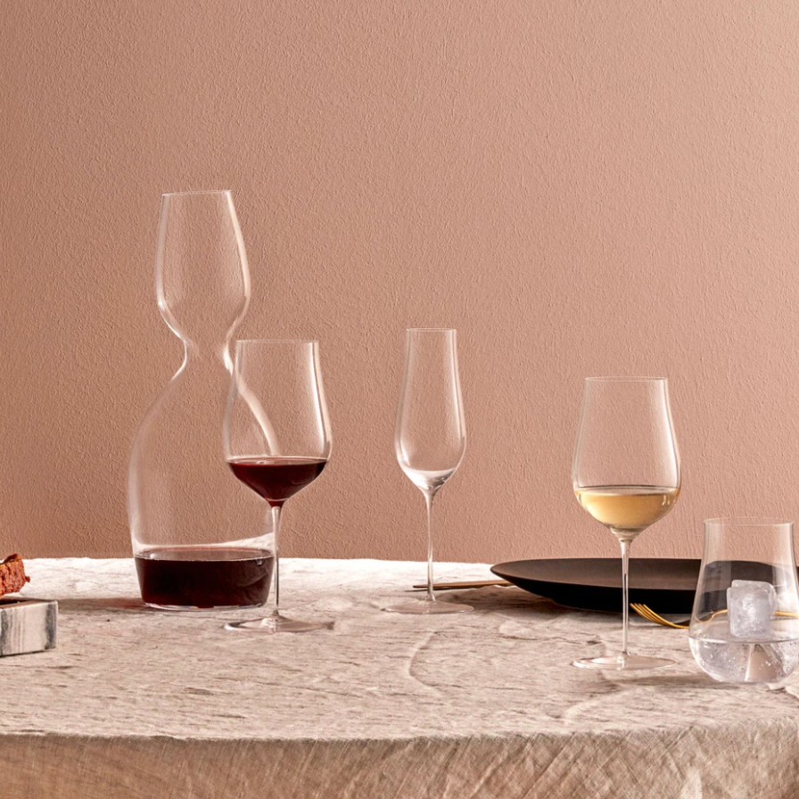 A Ghost Zero Tulip glass by Nude Glassware filled with red wine, a matching glass with white wine, a decanter, an iced water glass, a black plate, and bread rest on a beige tablecloth before a light brown wall.