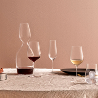 A Ghost Zero Tulip glass by Nude Glassware filled with red wine, a matching glass with white wine, a decanter, an iced water glass, a black plate, and bread rest on a beige tablecloth before a light brown wall.