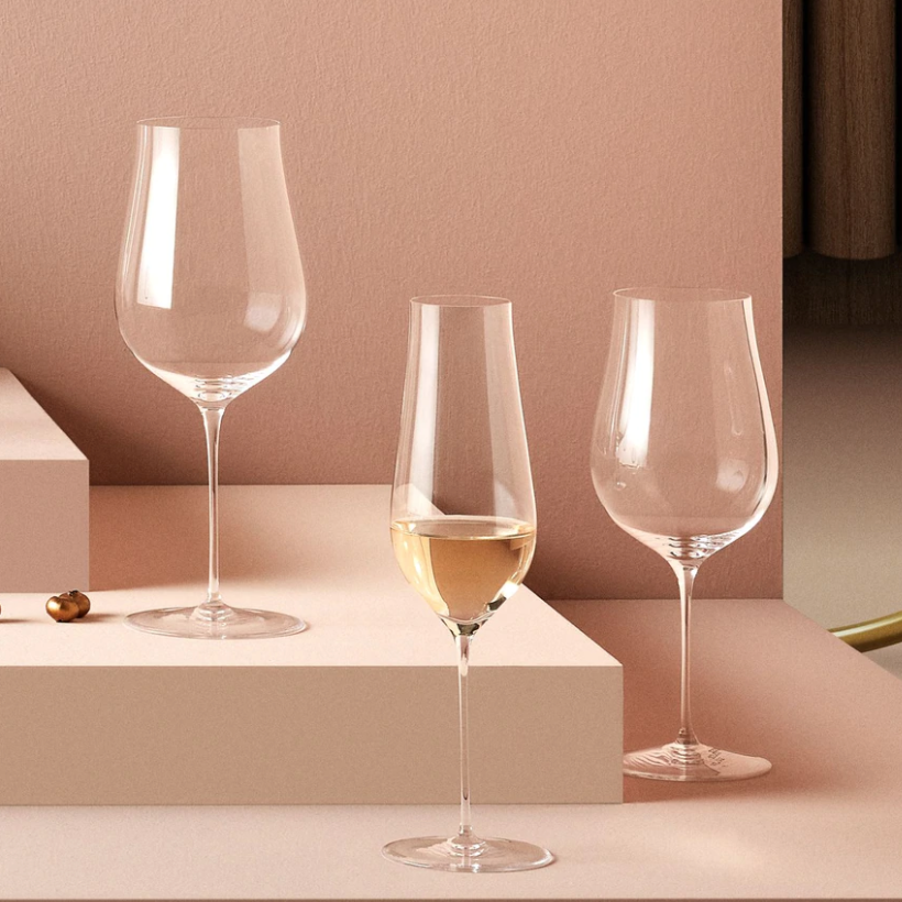 Three Ghost Zero Tulip Glasses by Nude Glassware, made from lead-free crystal, are displayed on a pink surface. One glass holds white wine, while two are empty. All have clear bowls and slender stems.