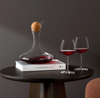 A dark round table holds a wine decanter with a cork stopper, set on The Kinfolk Home, and two Nude Glassware Mirage Barware lead-free crystal red wine glasses, all placed against a dark gray wall.