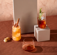 Three cocktails rest on a brown surface with beige blocks: one in a Nude Glassware Mirage Barware stemless glass with cinnamon and froth, another garnished with rosemary and ice, plus a plain brown drink. All glasses are lead-free crystal. Ginger root nearby.