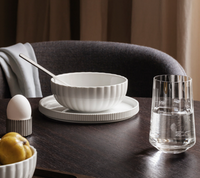A white ceramic bowl with a spoon rests on a matching plate atop a dark wood table, beside a Georg Jensen Bernadotte Glassware Set of 6, an egg in a holder, and yellow fruit. A gray chair and draped fabric complete the Scandinavian look.
