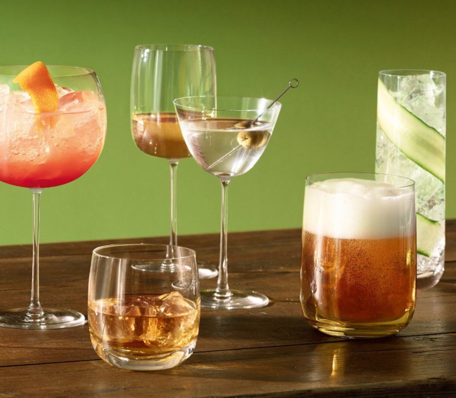 Six cocktails in clear, amber, pink, and foamy hues—garnished with items like cucumber and olive—are served in LSA’s Borough Barware Set of 4 glasses, all arranged on a wooden table against a green background.
