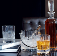 A crystal decanter with amber liquor sits beside a Baccarat Harmonie Barware Set of 2—one glass empty, one filled—plus an ice bucket with tongs and ice, all on a glossy table with books in the background.