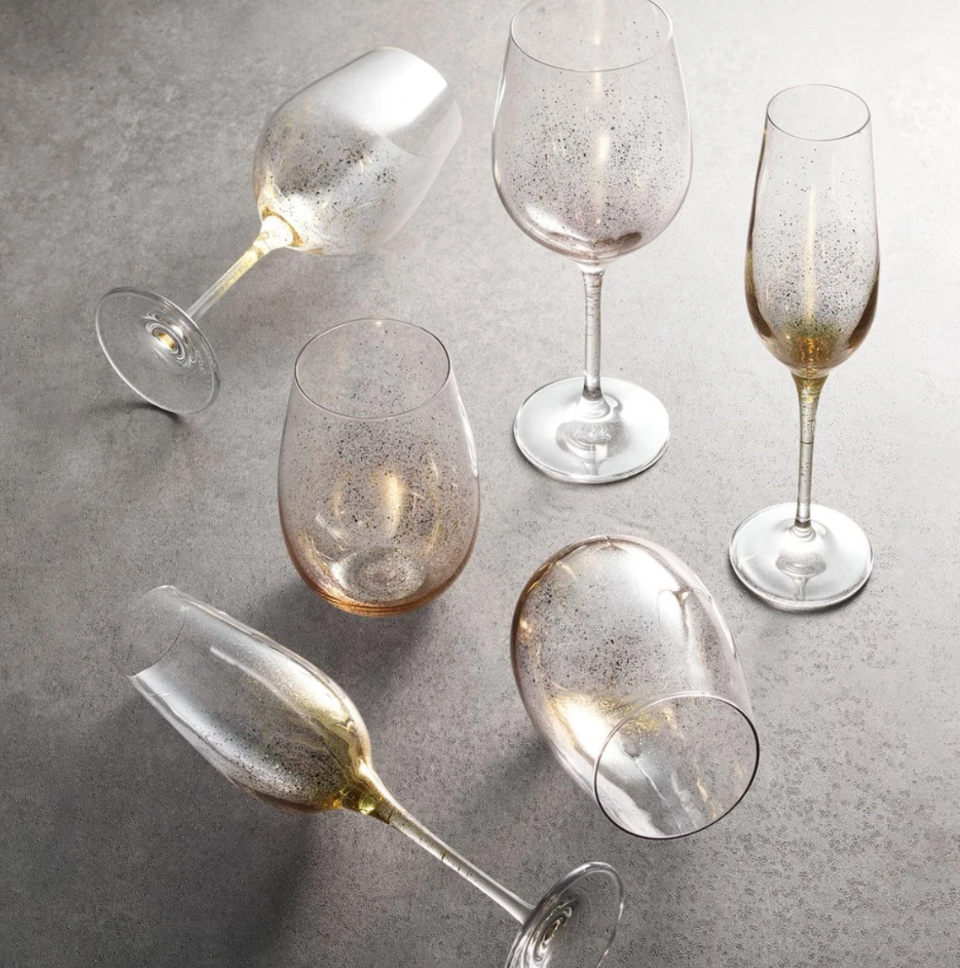 Four wine and champagne glasses from the Kim Seybert Orion Gold Barware Set are displayed on a gray surface, some upright and some on their sides, each featuring a hand-sprayed gold pattern inside for subtle elegance.