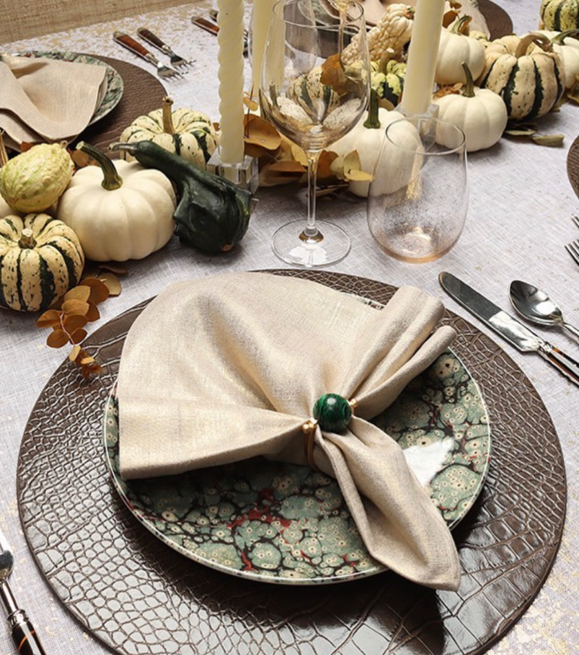 A festive table features patterned plates, a beige napkin with a green ring, candles, Kim Seybert’s Orion Gold Barware Set of 4, and a centerpiece of assorted gourds and pumpkins on a textured placemat.