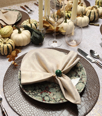 A festive table features patterned plates, a beige napkin with a green ring, candles, Kim Seybert’s Orion Gold Barware Set of 4, and a centerpiece of assorted gourds and pumpkins on a textured placemat.