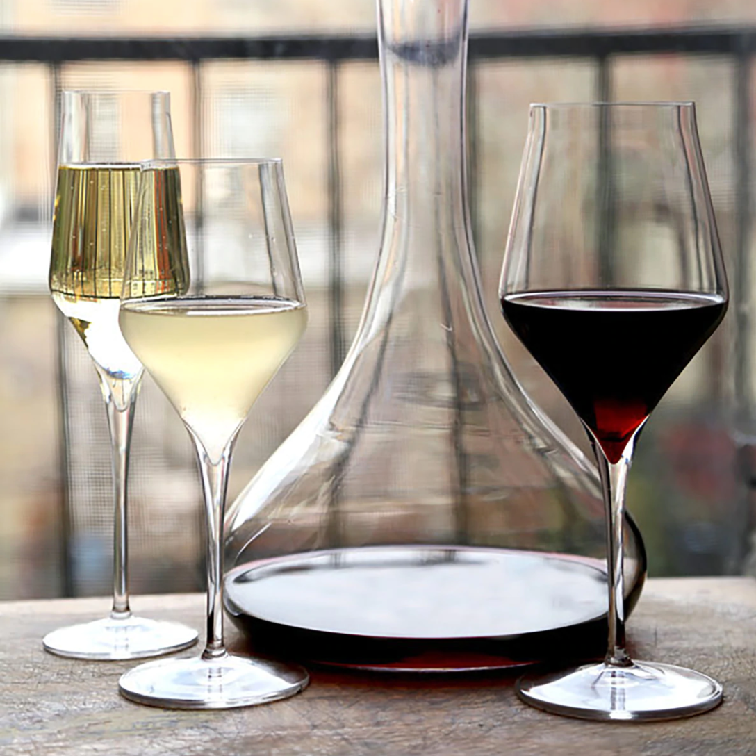 Two elegant Luigi Bormioli Supremo glasses, displayed on a wooden surface beside a decanter of red wine, stand out against a softly blurred background.