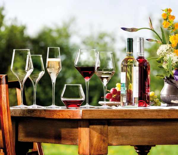 On a wooden table outdoors, two Luigi Bormioli Supremo glasses filled with red and white wine are displayed alongside bottles, grapes, and flowers; lush greenery forms the blurred background.