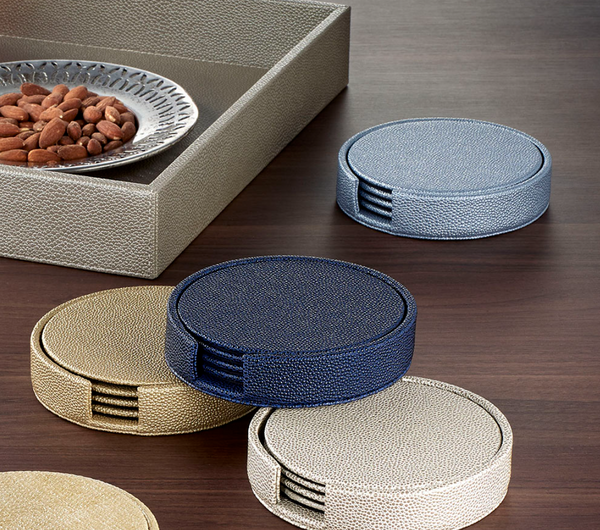 The Bodrum Skate Coaster Set of 4 features round, textured, phthalate-free vinyl coasters in blue, beige, and gold with a holder, neatly stacked on a wooden table next to a tray with almonds for added style and function.