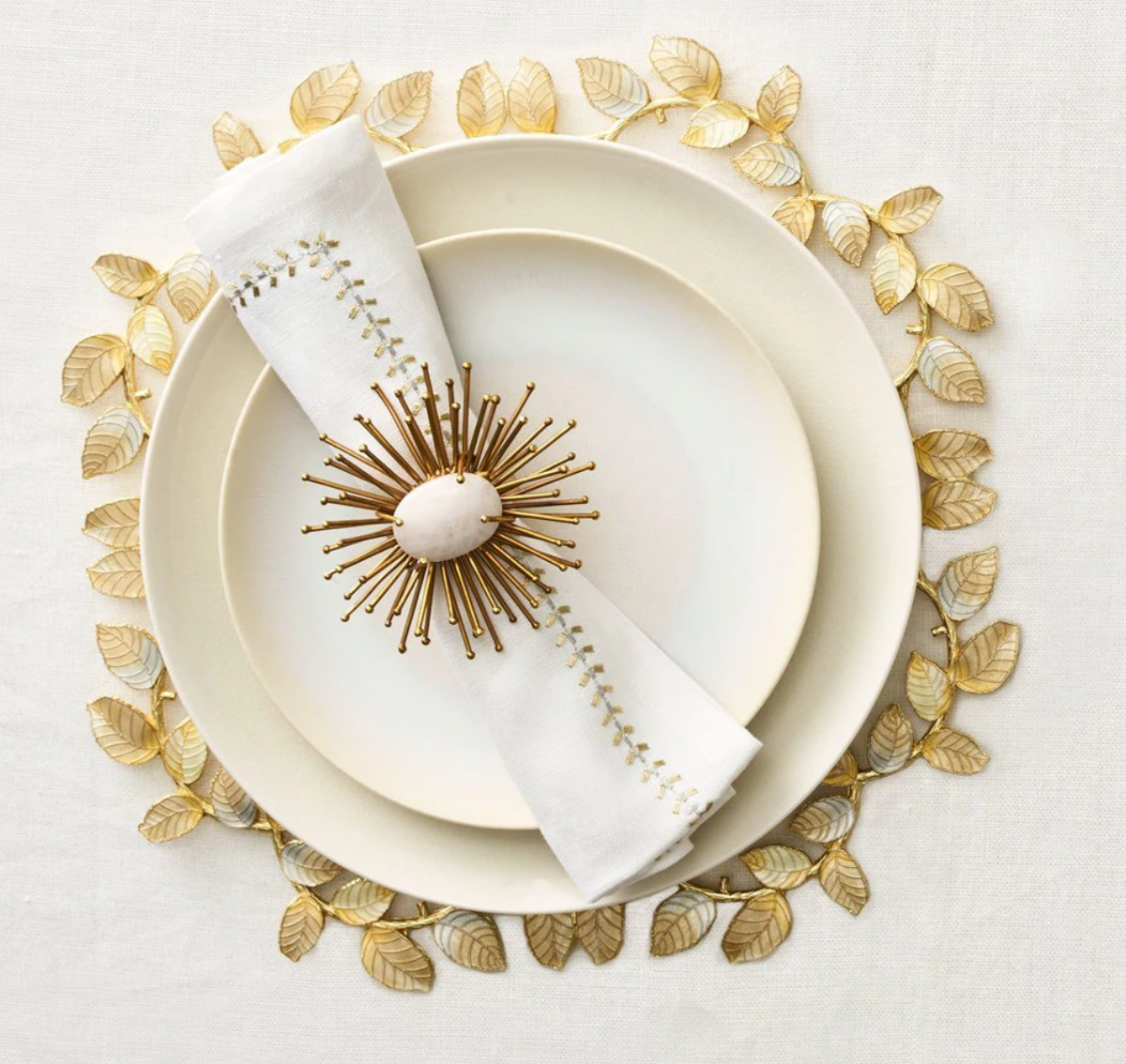 A white plate set with a gold leaf-shaped charger, a Kim Seybert Herringbone Napkin Gold & Silver, and a decorative gold sunburst napkin ring, arranged on a light tablecloth.