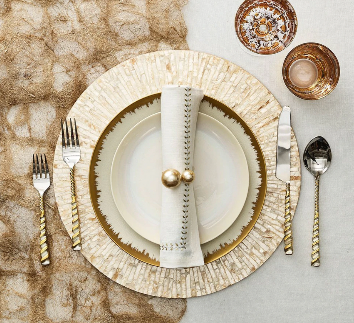 An elegant table features gold-accented cutlery, a cream and gold plate set, Kim Seybert Herringbone Napkin Gold & Silver Set of 4 with gold rings, and two gold-patterned glasses on a textured gold and beige tablecloth.