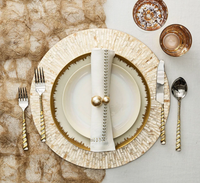 An elegant table features gold-accented cutlery, a cream and gold plate set, Kim Seybert Herringbone Napkin Gold & Silver Set of 4 with gold rings, and two gold-patterned glasses on a textured gold and beige tablecloth.