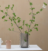 The Tina Frey Loopy Resin Vase Black, featuring decorative loops, holds tall green branches and a single white flower. This large vase sits on a white surface next to two small ceramic vessels against a beige background.