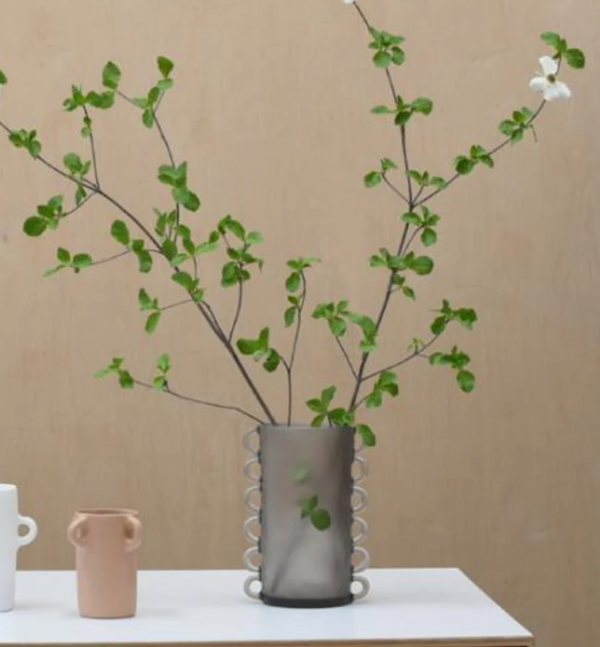 The Tina Frey Loopy Resin Vase Black, featuring decorative loops, holds tall green branches and a single white flower. This large vase sits on a white surface next to two small ceramic vessels against a beige background.
