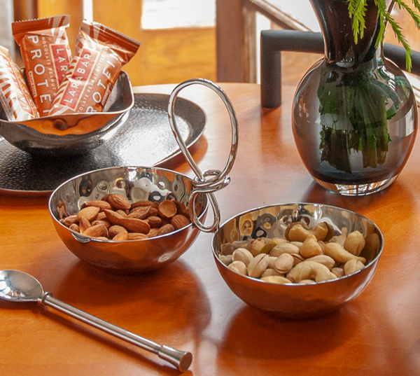 Two metal Opus Two Bowl Dishes by Mary Jurek rest on a wooden table, one with almonds, the other with cashews. A nearby spoon, protein bars, and a vase of greenery complete this scene—an ideal housewarming gift.