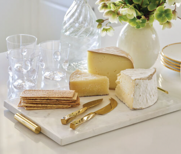 The Aerin Leon Gold Cheese Set includes three cheeses, crackers, brass-plated cheese knives, two clear glasses, a textured glass bottle, and a vase of green flowers on a white table.