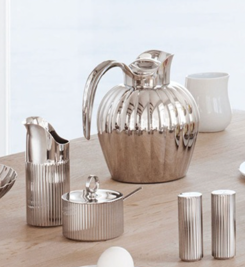 A set of shiny silver tableware, featuring the Georg Jensen Bernadotte Salt & Pepper Set, sits on a light wooden table with a white ceramic cup in the background.