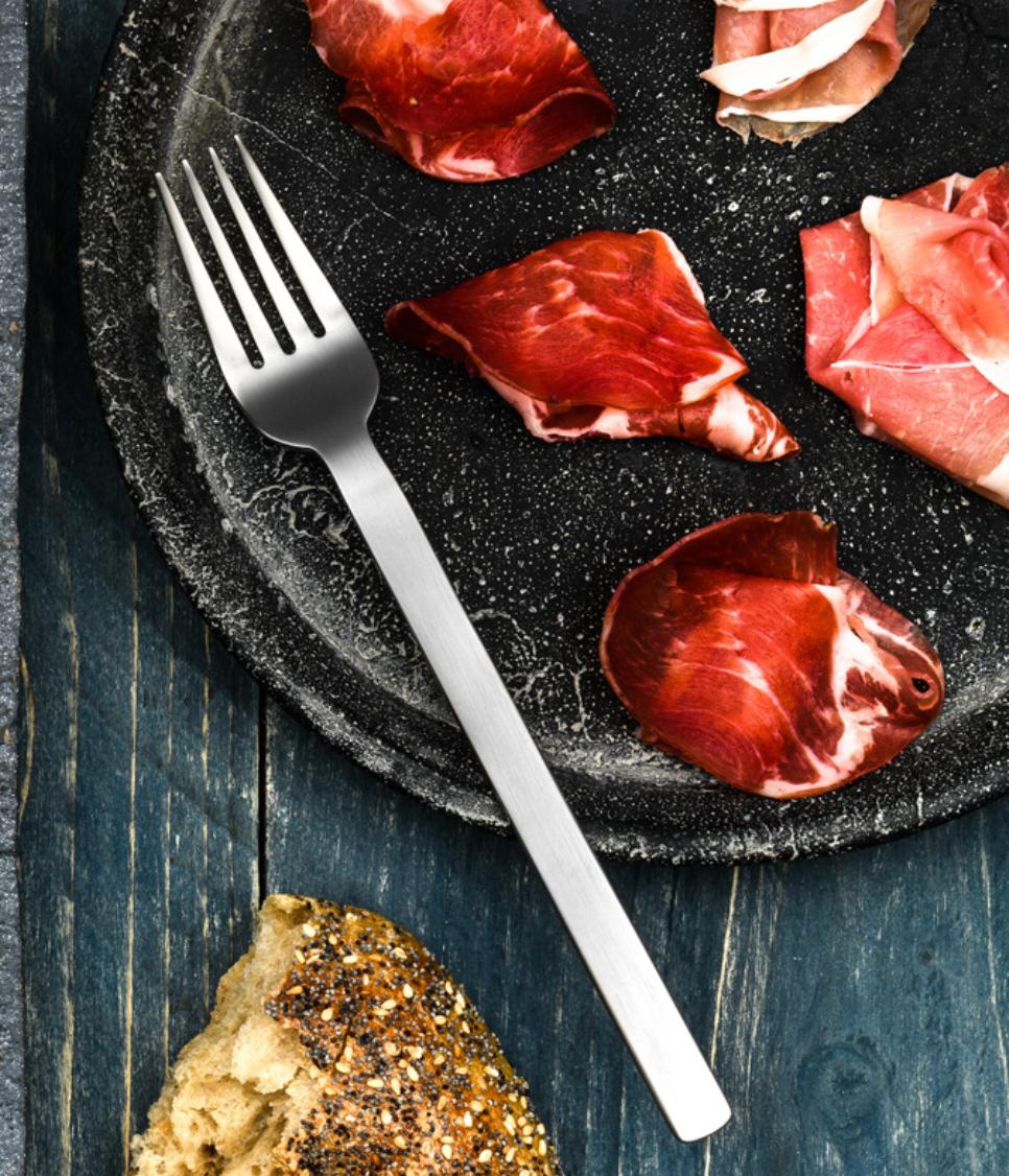 Close-up of thinly sliced cured meat on a dark plate with a Mepra Stile Ice Flatware 5 Piece Setting silver fork, made from 18/10 stainless steel. A seeded bread slice rests on the rustic wooden surface beside the plate.