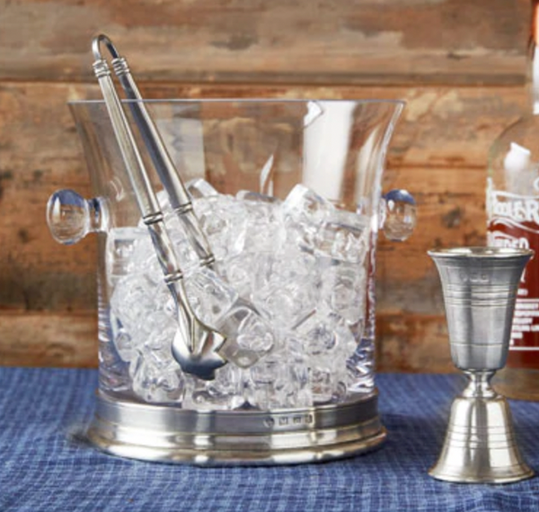 The MATCH Pewter & Crystal Ice Bucket With Handles by MATCH, filled with ice and silver tongs, sits beside a jigger and bottle on a blue cloth—handmade Italian barware set against a warm wooden backdrop.