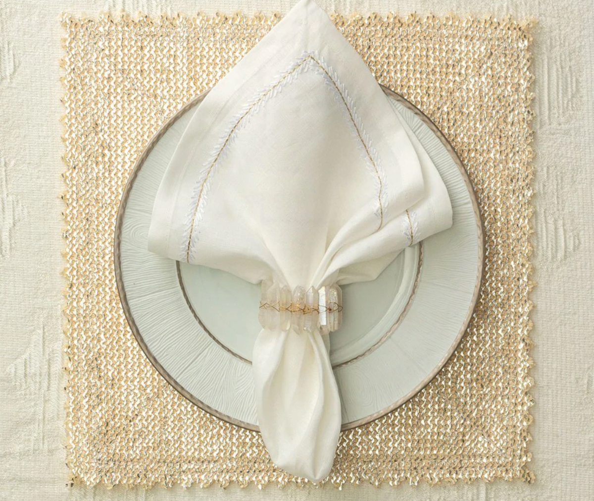 A white cloth napkin with a Kim Seybert Radiant Napkin Ring Iridescent is neatly placed on a white plate with silver edges, resting on a gold woven placemat atop a cream-colored tablecloth.
